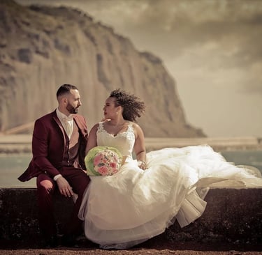 Mariés assis sur muret face à la mer et falaise, costume bordeaux et robe princesse, bouquet pastel.
