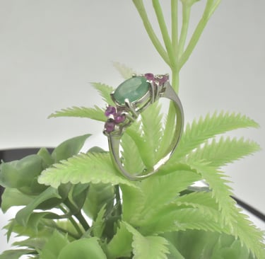 Elegant silver ring featuring an oval green emerald and small ruby side stones on a green plant.