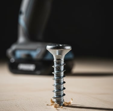 Close-up of a silver metal screw driven into a wood plank with a cordless power drill in the background.