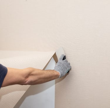 A worker's gloved hand carefully peeling or hanging textured beige wallpaper on a wall during home renovation.