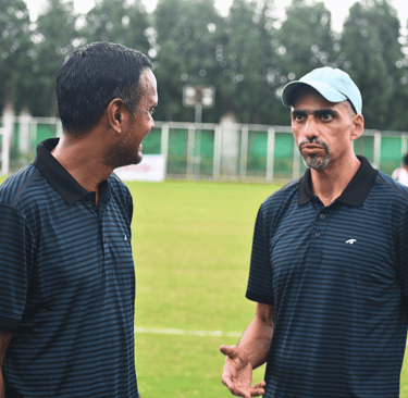 Two soccer coaches in striped polo shirts discuss strategy on a green football field.