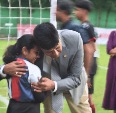 A smiling man in a suit hugging a young female soccer player on a green football field.