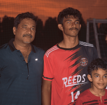 A soccer player in a Reeds Football Club jersey poses with two family members at sunset.