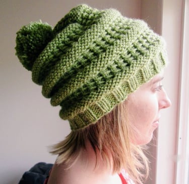 A young woman staring out a window wearing a handknit green slouchy beanie with a pompom