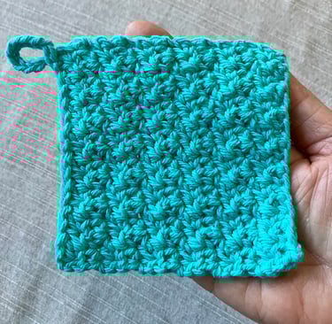A hand holding a handmade crocheted aqua face scrubby with textured stitch