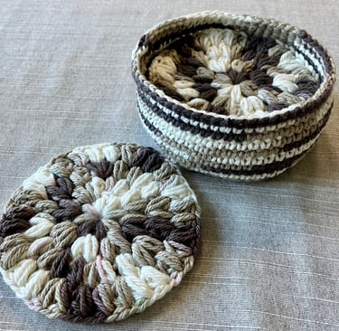 Crocheted brown, tan, and cream colored drink coaster with more of the same coasters inside a basket