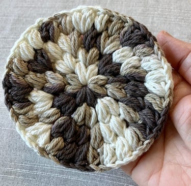 A hand holding a brown, tan, and cream colored crocheted drink coaster.