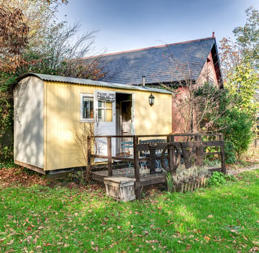 A rustic yellow shepherd's hut with a wooden deck and table set in a green garden.