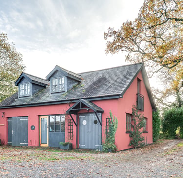 A red cottage with grey shutters and dormer windows nestled among autumn trees and fallen leaves.