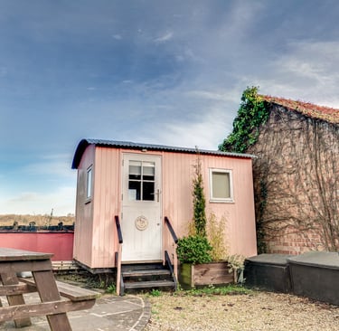 A pink shepherd's hut with a wooden picnic table and rustic brick wall.