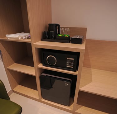 Hotel room amenities featuring a digital safe, mini fridge, and electric kettle on wooden shelves.