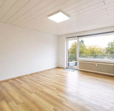 Leeres, modernes Wohnzimmer mit hellem Laminatboden und einem großen Fenster mit Blick auf einen Balkon.