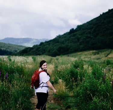 Smiling woman with a red backpack hiking on a scenic mountain trail through a lush green valley.