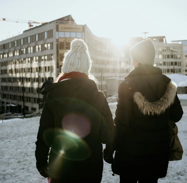 Two women in winter coats and beanies walking through a snowy city at sunset.