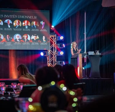 A woman speaking at a gala podium next to a digital screen showing guests of honor.