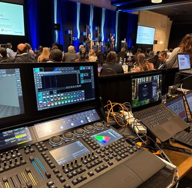 Professional lighting console and laptops at a live event production tech booth in a conference hall.