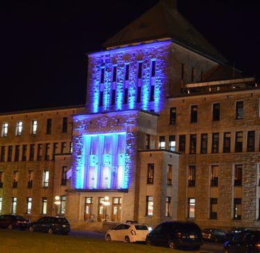 Large stone building with many windows, illuminated by blue lights at night, with cars parked along the street.