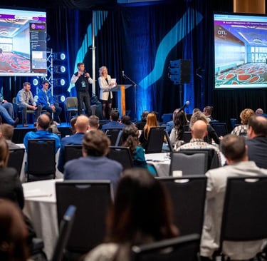Speakers present at a corporate conference panel on stage with large digital screens for the audience.
