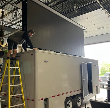A technician installs a large mobile LED screen on a white custom trailer inside a warehouse.