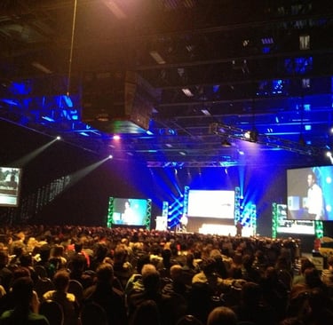Crowd at a large tech conference with blue stage lighting and large video screens.