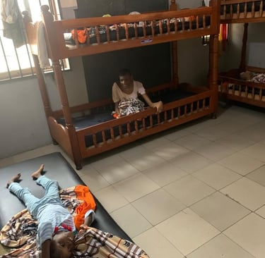 A person sitting on a wooden bunk bed and a child resting on a floor mattress in a shared dormitory room.