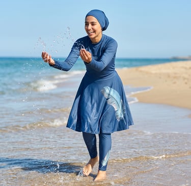 A woman wearing a blue modest burkini swimsuit with a feather print splashes water at the beach.