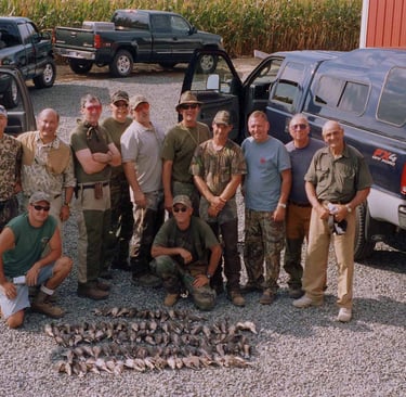 A large group of hunters in outdoor gear posing with harvested doves in a gravel lot next to several pickup trucks.