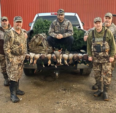 Five hunters in camouflage gear posing with harvested ducks on the tailgate of a white pickup truck.