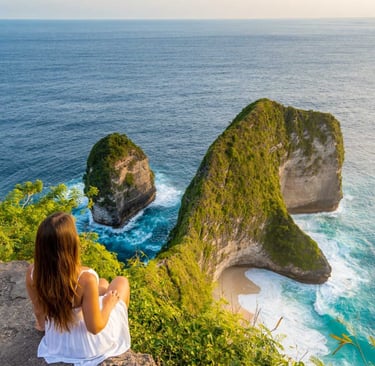 A woman in a white dress sits on a cliff overlooking Kelingking Beach in Nusa Penida, Bali.