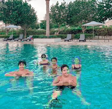A group of happy children smiling and swimming in a resort swimming pool with lounge chairs and palm trees.