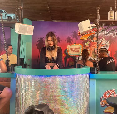 Kat Bendis performing at a vibrant Ibiza beach club bar with tropical decor and neon signs.