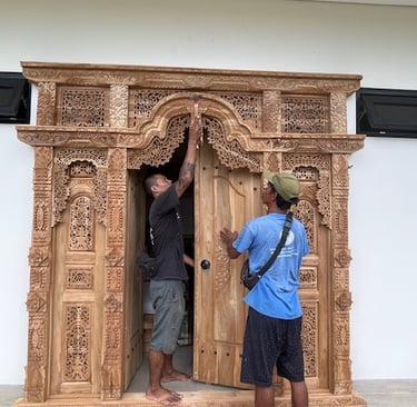 Two craftsmen installing a hand-carved teak wood Balinese door with intricate floral patterns.