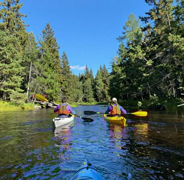 Two kayakers paddling through crystal-clear water framed by lush green forest under a bright blue sky.