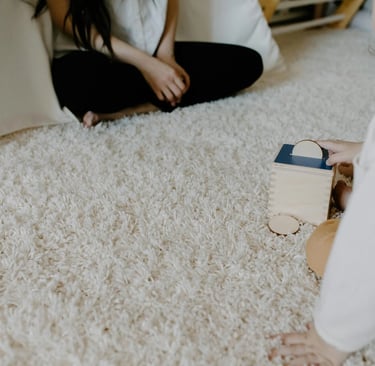 A toddler plays with a wooden Montessori coin box toy on a plush rug near a play tent.