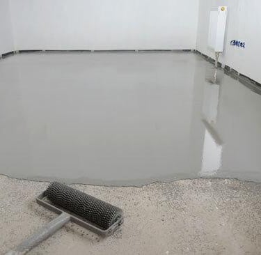 A gray self-leveling epoxy floor coating being applied with a spiked roller in a room.