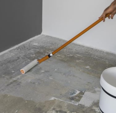 A person applying a clear concrete primer to a garage floor using a long-handled paint roller.