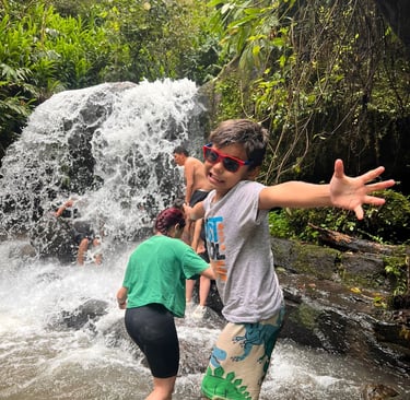 Un niño con gafas de sol posa emocionado frente a una cascada en la selva tropical junto a su famili
