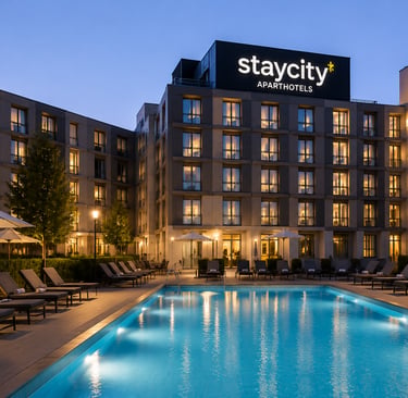 Staycity Aparthotels building at night with a lit swimming pool and outdoor lounge chairs.