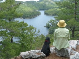 Marlies siiting on rocks overlooking lake with black dog by her side.