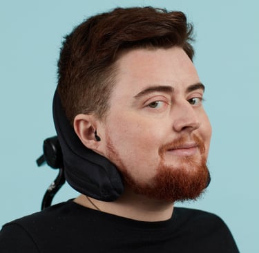 Jamie, a white person on a blue background, electric wheelchair headrest behind their head.