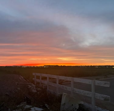 A vibrant orange and purple sunset over a dark field with a white wooden fence in the foreground.