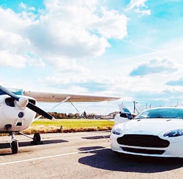 A white Aston Martin luxury sports car parked next to a small private propeller plane on an airfield runway.