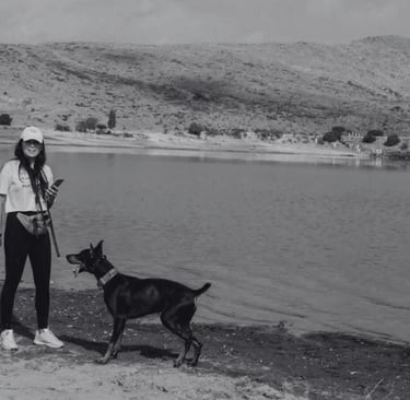 Una mujer junto a su Doberman Pinscher negro en la orilla de un lago con colinas al fondo.