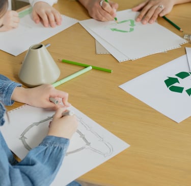 school children sitting at a table drawing recycling symbols in green pencil.