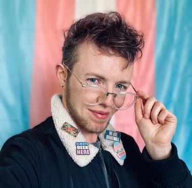 White trans masc in front of trans Pride flag wears a jacket with trans visibility pin badges