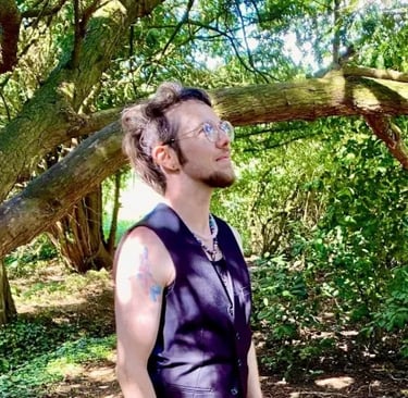 William Elisabeth Cuthbert, a white trans masc person, with trees in the background