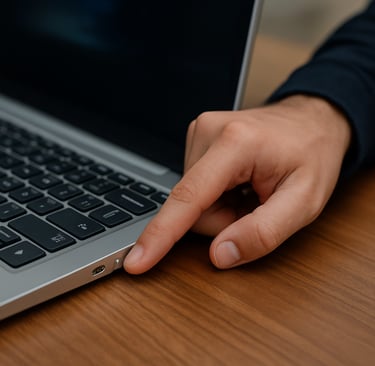 Pessoa pressionando o botão de ligar de um notebook prateado sobre uma mesa de madeira, em um ambien