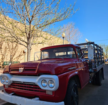Classic red Ford flatbed truck parked on a sunny city street with budding spring trees.