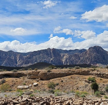 Panoramic view of rugged desert mountains and rock formations under a bright blue sky with white clouds.