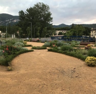 A scenic garden park with sandy paths, lush flowering plants, a blue bridge near Quillan mountains.